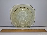 Top view of yellow square Depression glass plate showing detailed embossing and scalloped corners with a ruler to demonstrate size (9 inches).
