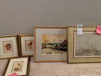 Wide view showing all framed pieces: two smaller pressed flower artworks in gold frames, one pressed flower artwork in brown frame, larger colorful landscape painting in wood frame, and monochromatic etching in gold frame.