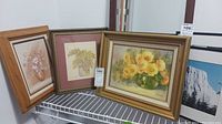 Three framed flower paintings displayed together, one smaller with neutral colored flowers, two larger with yellow flowers in green vases.