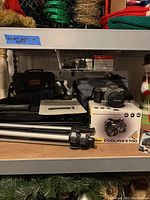 Shelf containing Canon camera bag, manual, Denon CD player, Nikon Coolpix 5700 box with camera on top, and tripod.