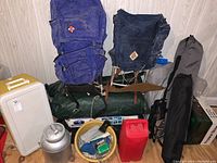 Photo showing two large blue backpacks, insulated cooler, metal milk canister, green dining canopy bag, red container, yellow basin with hatchet and camping supplies, and additional camping gear.