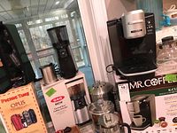 Photo showing Mr. Coffee single cup brewing system placed on its original box, OXO Brew coffee grinder on its box next to a stainless steel French press and an ice bucket.