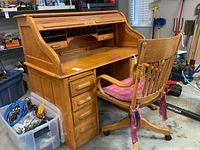 Full view of solid oak roll top desk placed in garage with swivel chair in front. Shows desk's curved roll top open and the multiple drawers and cubbies.