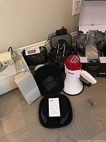 Various electronic items including white speakers, Bluetooth speaker in box, carrying case, and megaphone arranged on wooden floor.