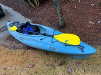 Full view of blue kayak on grass with paddle across cockpit and life jacket on seat