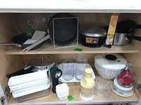 View of kitchen shelf with various kitchen items including plastic storage containers, glass jars, salad spinner, flashlight, chopper, glass cups, and plastic kitchen tools.