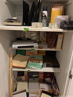Shelves inside closet with laminating sheets, copy paper, file folders, envelopes, adding machine, tape rolls, and various office papers.