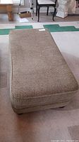 Full view of rectangular ottoman upholstered in brown tweed fabric, placed on a carpeted floor.