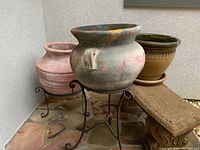 Three large pottery planters positioned outdoors. Two supported by metal stands, one with matching saucer.