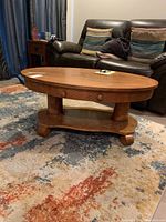Side and top view of oval quartersawn oak cocktail table on multi-colored rug with dark leather couch in background.