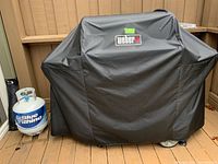 Weber grill covered with black Weber branded protective cover on wooden porch deck with Blue Rhino propane tank next to it.