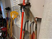 Photo showing several yard tools hanging on wall including shovels, rakes, hoes, pitchfork, and a bright orange pole pruning saw.