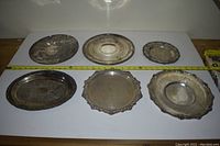 Top down photo of six assorted silver plated platters arranged in two rows, showing front sides with tarnishing.