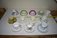 Full view of eleven assorted teacups and matching saucers arranged in two rows, along with one white ceramic flower-shaped item to the side.