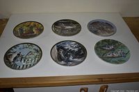 Six decorative collector plates laid out on a surface showing wildlife scenes with birds, cats, and squirrels in nature settings.