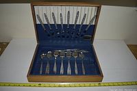 Full view of the cutlery set displayed in an open wooden box with blue velvet interior, showing knives on the lid, and spoons, forks, and serving utensils in the base.