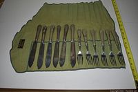 Full view of 12 piece fish cutlery set rolled out on white surface, includes 6 knives and 6 forks, wooden handles and tarnished silver blades/forks, on a green fabric holder.