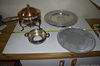 Image showing copper fondue pot with wooden handle and brass lid, two serving platters—one embossed and one plain—and small warming bowl with two handles, all placed on a white surface.