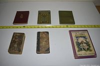 Six old books displayed on a surface with a measuring tape showing length approximately 12 inches. Books exhibit wear and damage; one book cover missing front, side and back covers, another book colorful with illustrated cover.