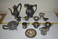 View of entire set showing large water jug, teapot, sugar and cream dishes and other silver serving pieces on white surface with measuring tape.