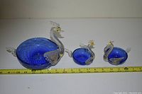Photo of three glass swan figurines on a white surface with measuring tape indicating sizes.