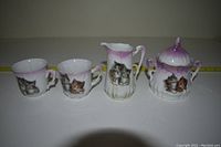 Front view of the small cat-themed porcelain tea set showing two cups, creamer, and sugar bowl with kitten images and purple tops.