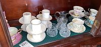 Overall view of assorted teacups, saucers, and crystal tableware arranged on a wooden surface with glass cabinet background.