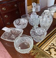 Group of nine crystal items including three decanters with stoppers, various bowls, and vases on a wooden table.