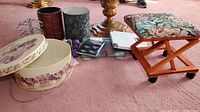 Photo shows floral upholstered rolling footstool, decorative round hat boxes, gold decorative stands, digital scale, hand weights and various items on carpeted floor.