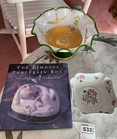 Top down view of large glass fruit bowl, Limoges coffee table book, and two Limoges porcelain dishes on marble table.