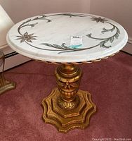 Front angled view of round marble top table showing floral inlays and ornate gold painted wooden pedestal base.