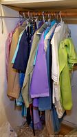 Photo of assorted women's clothing hanging in closet showing jackets, blouses and tops in various pastel and neutral colors, photographed from front side