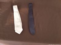 Photo showing two men's neckties laid flat on a brown surface, one white and one dark navy blue.