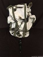 White backpack with black camouflage-like pattern and green straps lying against a black background with Infantry watch placed in front.