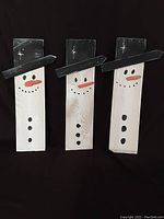 Three wooden snowmen decorations placed side-by-side on a black background showing their front painted faces and hats.