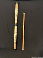 Wooden didgeridoo and thinner wooden rainstick shown side by side against dark backdrop.