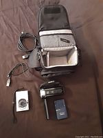 Canon HD video camera, Canon digital camera, camera case, and various cables arranged on a brown fabric surface. The camera case is open showing padded compartments and mesh pockets.
