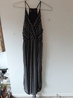 Full view of a sleeveless black and white geometric patterned dress with thin shoulder straps hung on a hanger against a white wall.