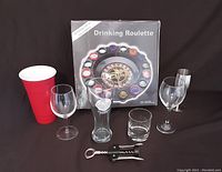 Photo showing a red plastic cup, five other different types of drinking glasses, a metal shot glass, corkscrew, and boxed Drinking Roulette game set