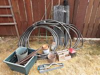 Photo showing all gardening supplies including coiled hose, pots, fencing, brush, stakes, and planter box on grass against wooden fence background.