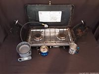 Front view of two-burner camping gas stove with cooking grates, attached fuel hose and valve, alongside small pans and mugs for outdoor cooking.