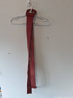 Red belt hanging on wall-mounted hook. Shows solid red smooth side and opposite side with textured pattern. Adjustable holes visible. Red rectangular buckle present.