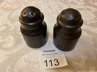 Front view of two black salt and pepper shakers placed on a floral patterned cloth with a MaxSold lot tag showing number 113.