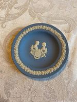 Top-down view of the round blue jasperware style Wedgewood plate with white relief profiles and floral border on a cream patterned fabric surface.