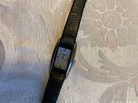 Photo showing rectangular stainless steel case watch with white face and black leather strap laid flat on surface.