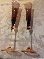 Two handmade champagne flutes shown upright on a table with a textured beige tablecloth, displaying their purple to pink gradient coloring and clear stems.