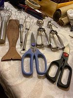Photo showing three metal whisks, two metal ice cream scoops, one wooden whale tail spatula, and two pairs of kitchen scissors on a table.