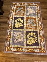 Full view of rectangular handmade prayer rug showing six floral panels with a floral border on wooden floor