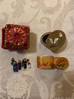 Top view of three small boxes on fabric surface: red sequinned square box, round soapstone box with mother of pearl lid, small wooden painted box with miniature worry dolls.
