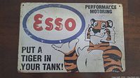 Photo of a 12x8 inch Esso metal sign with tiger mascot, slogan, and drilled mounting holes at corners.